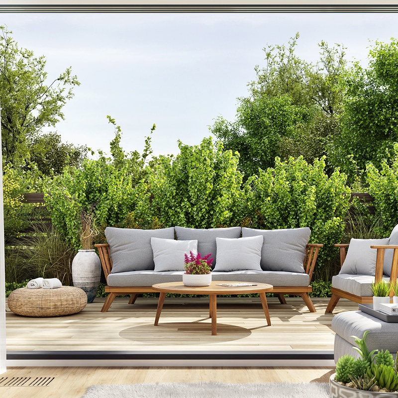 Salon de jardin moderne sur terrasse en bois clair, canapé gris et table basse devant une haie de verdure luxuriante.